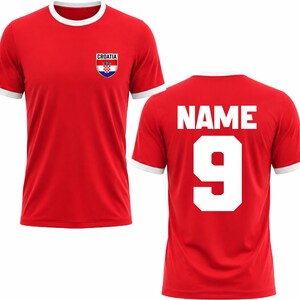 Puede incluir: Camiseta roja con ribetes blancos en el cuello y las mangas. La parte delantera presenta un emblema de Croacia y la palabra "CROATIA". La parte trasera muestra la palabra "NAME" sobre el n&uacute;mero "9" en blanco.