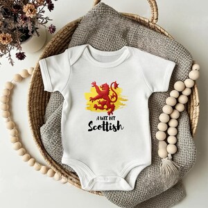 May include: White baby onesie with a red lion emblem on a yellow background. The text "A wee bit Scottish" is printed below the emblem. The onesie is in a woven basket with a gray blanket.
