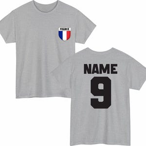 May include: A heather gray t-shirt with a French flag emblem on the front and the word "NAME" and the number "9" on the back in black. The shirt has a crew neck and short sleeves.