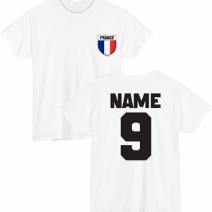 May include: White t-shirt with a French flag emblem on the front and the word "NAME" above the number "9" on the back. The emblem is a shield shape with the colors blue, white, and red. The number 9 is in a bold, black font.