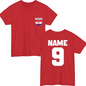 May include: A red t-shirt featuring a Croatian flag emblem on the front and the number 9 with the word "NAME" on the back. The emblem is a shield shape with the Croatian flag design. The shirt is a short-sleeved crew neck.