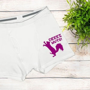 May include: White boxer briefs featuring a dark pink graphic of a squirrel and the text DEEEZ NUTS! printed on the right leg. The underwear has a grey elastic waistband with a centered grey tab detail and visible stitching along the seams.