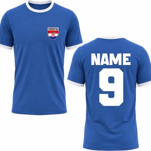 Puede incluir: Una camiseta azul con ribetes blancos en el cuello y las mangas. La parte delantera presenta un emblema de Croacia y la palabra "CROATIA". La parte trasera muestra el n&uacute;mero "9" y la palabra "NAME" en blanco.