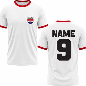 Puede incluir: Camiseta blanca con ribetes rojos en el cuello y las mangas. La parte delantera presenta un emblema de la bandera croata y la palabra "CROATIA". La parte trasera muestra el n&uacute;mero "9" y la palabra "NAME" en negro.