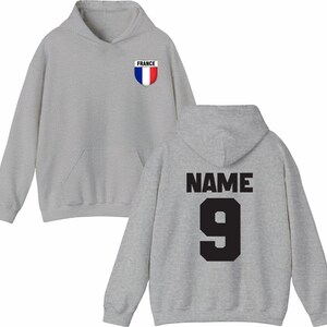 May include: A light gray hoodie with a front pocket and a hood. The front features a small emblem with the word "FRANCE" and a red, white, and blue shield. The back displays the word "NAME" above the number "9" in black.