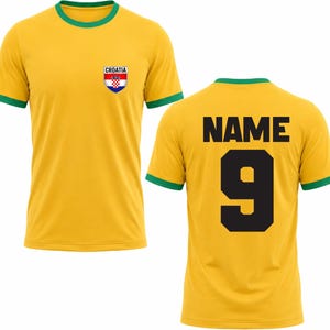 Puede incluir: Una camiseta de f&uacute;tbol amarilla con ribetes verdes y un escudo croata en el pecho. La parte trasera muestra el n&uacute;mero 9 y la palabra "NAME" en negro. La camiseta est&aacute; hecha de un material ligero.