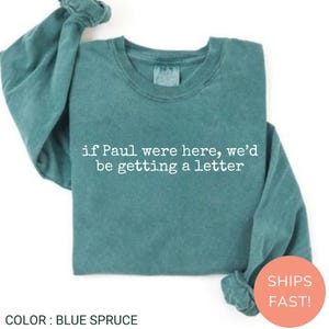 May include: A blue spruce long-sleeve crewneck sweatshirt featuring white text printed on the front that reads, if Paul were here, we'd be getting a letter. The garment has a ribbed collar, cuffs, and hem with a soft, washed-out fabric texture.