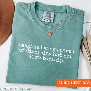 May include: Seafoam green t-shirt with the text "imagine being scared of diversity but not dictatorship" in white. The shirt has a crew neck and short sleeves. A silver necklace with a pendant is draped on the shirt.