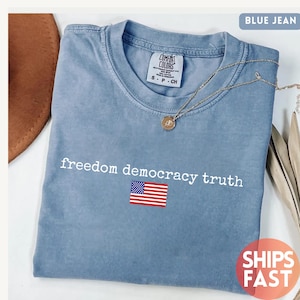 Könnte beinhalten: Ein jeansblaues T-Shirt mit Rundhalsausschnitt und einem Etikett der Marke Comfort Colors. Auf der Vorderseite befindet sich der weiße Schriftzug "freedom democracy truth" über einer kleinen Grafik der Flagge der Vereinigten Staaten. Eine goldene Kette liegt auf dem Stoff.
