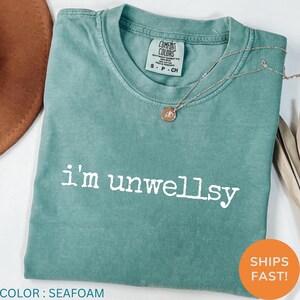 May include: A seafoam green short-sleeve t-shirt with a crew neckline and the white text "i'm unwellsy" printed across the chest. The shirt features a Comfort Colors brand label at the inner neck and is paired with a thin silver-toned necklace.