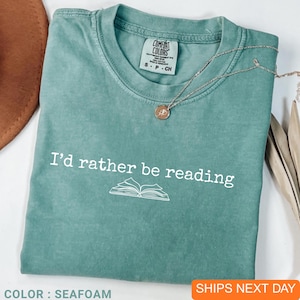 May include: Seafoam green t-shirt with the text "I'd rather be reading" and an open book graphic. The shirt is a Comfort Colors brand, size S-P-CH. A silver necklace with a small pendant is draped across the shirt.