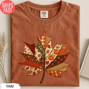 Peut inclure: Un t-shirt à col rond couleur rouille arborant un grand motif central en forme de feuille d'érable, composé de divers motifs de tissus style patchwork dans des tons automnaux, incluant des carreaux, des fleurs et des imprimés unis avec deux citrouilles.