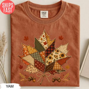 May include: A rust-colored crew neck t-shirt featuring a large central graphic of a maple leaf composed of various patchwork fabric patterns including sunflowers, plaid, and floral prints, with two small acorns and wheat stalks at the base.