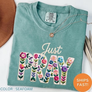 May include: Seafoam green t-shirt with the embroidered phrase "Just Pray" decorated with colorful floral designs. The shirt has a crew neck and short sleeves. A silver necklace with a pendant is also visible.