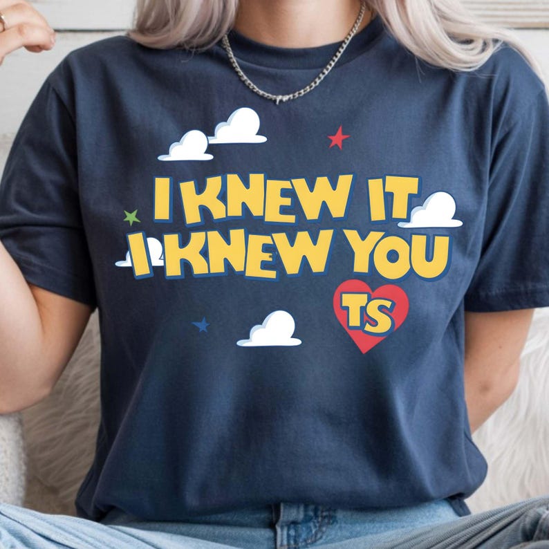 Puede incluir: Una camiseta de manga corta azul marino oscuro con el texto amarillo I KNEW IT I KNEW YOU TS en fuente negrita. El dise&ntilde;o incluye gr&aacute;ficos de nubes blancas, un coraz&oacute;n rojo y peque&ntilde;as estrellas azules, verdes y rojas en el pecho.