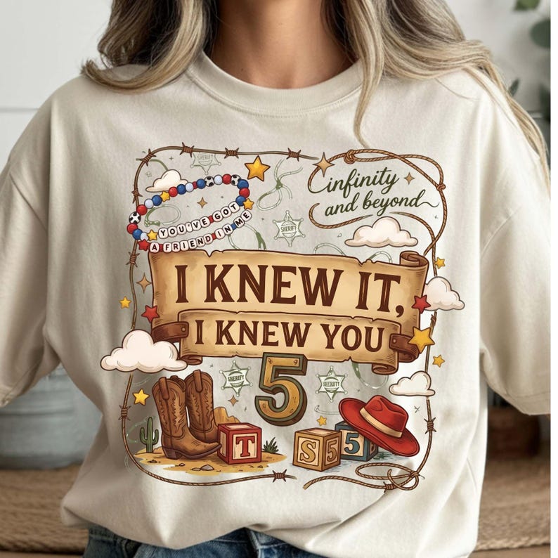 Puede incluir: Una camiseta color crema con un estampado gr&aacute;fico rodeado por un lazo, que incluye botas de vaquero, un sombrero rojo, bloques de letras y el texto "Cinfinity and beyond", "YOU'VE GOT A FRIEND IN ME" e "I KNEW IT, I KNEW YOU 5".