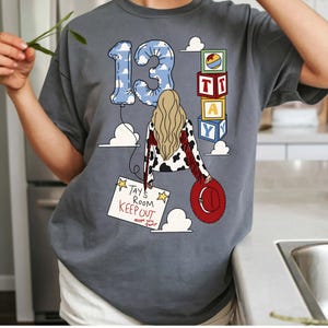 Puede incluir: Una camiseta gris carb&oacute;n con un gr&aacute;fico de una persona con cabello largo rubio, una chaqueta con estampado de vaca y un sombrero rojo. El dise&ntilde;o incluye un globo azul con nubes en forma de 13, bloques de juguete apilados con TAY y un letrero que dice TAY'S ROOM KEEP OUT.