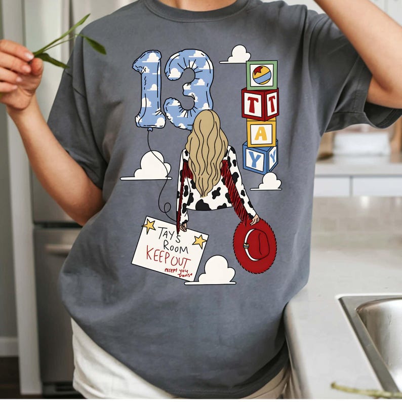 Puede incluir: Una camiseta gris carb&oacute;n con un gr&aacute;fico de una persona con cabello largo rubio, una chaqueta con estampado de vaca y un sombrero rojo. El dise&ntilde;o incluye un globo azul con nubes en forma de 13, bloques de juguete apilados con TAY y un letrero que dice TAY'S ROOM KEEP OUT.