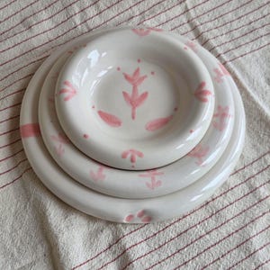 May include: Three white ceramic plates with a pink floral pattern, stacked together. The plates are of varying sizes, with the smallest at the top. The floral design features stylized leaves and dots.