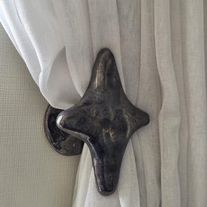 Puede incluir: Un alzapaños de cortina de metal oscuro con forma de estrella de cuatro puntas y una superficie texturizada e irregular. El objeto está montado en una pared y sujeta una cortina de tela blanca semitransparente fruncida, creando un contraste.