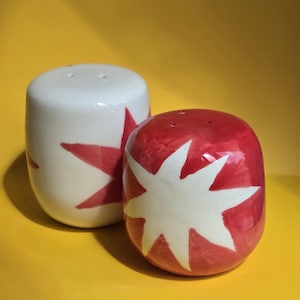 May include: A pair of ceramic salt and pepper shakers. One is white with a red star design, and the other is red with a white star design. Both have small holes on top for dispensing.