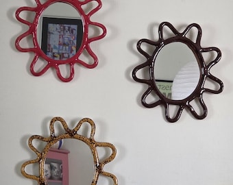 May include: Three wall mirrors with irregular, wavy, sun-shaped frames. One frame is glossy red, one is dark brown, and one is a speckled mustard yellow. Each mirror has a central circular glass pane reflecting the surrounding room.