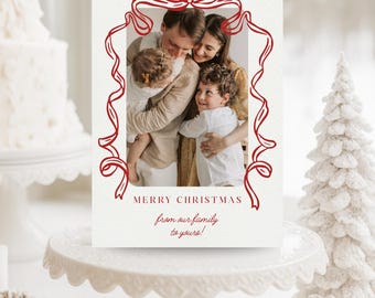 Könnte beinhalten: Eine Weihnachtskarte mit einem Familienfoto, das von einem roten Schleifendesign umrahmt ist. Auf der Karte steht "MERRY CHRISTMAS from our family to yours!" Sie wird auf einer weißen Tortenplatte mit einem dekorativen Weihnachtsbaum und einer Kiefernzapfen im Hintergrund präsentiert.
