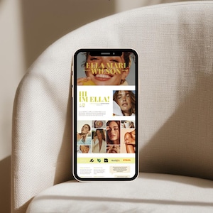 May include: A smartphone displays a website with the text "ELLA MARI WILSON" and "HI IM ELLA!" The website features multiple images of a woman, with a light yellow and white color scheme. The phone is resting on a beige armchair.