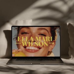 May include: A laptop displaying a close-up of a smiling woman holding a peach. The screen features the text "ELLA MARI WILSON CONTENT CREATOR" in a gold font. The laptop is on a white surface, with a soft, neutral background.