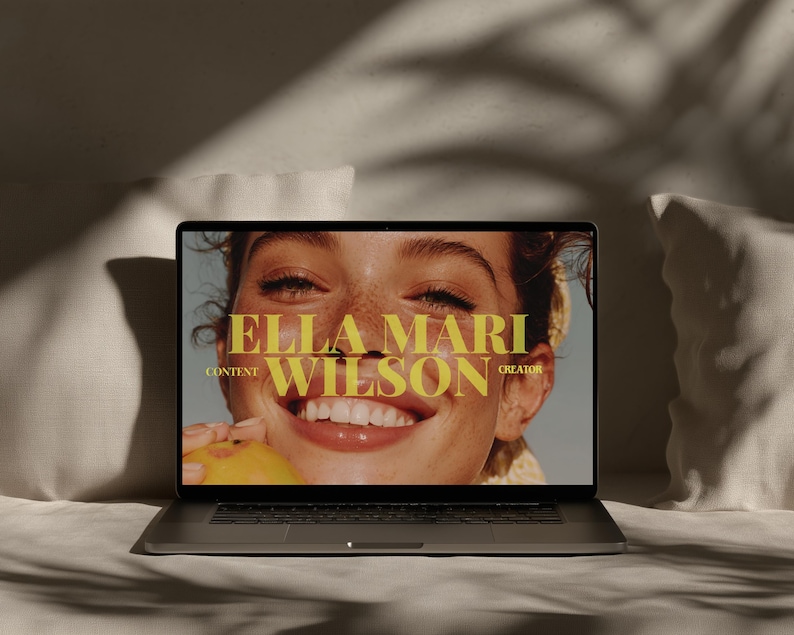 May include: A laptop displaying a close-up of a smiling woman holding a peach. The screen features the text "ELLA MARI WILSON CONTENT CREATOR" in a gold font. The laptop is on a white surface, with a soft, neutral background.