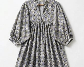 Puede incluir: Un vestido largo y fluido en un tono morado claro, adornado con un estampado floral repetido en tonos verdes y blancos. El vestido presenta un cuello alto con volantes, un escote en V con un panel a rayas y mangas voluminosas. El vestido está colgado en una percha de madera.