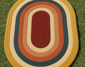 May include: Oval-shaped rug with a retro design. The rug features concentric oval bands in various colors, including yellow, cream, blue, orange, and burgundy. The rug is placed on a green grassy surface.