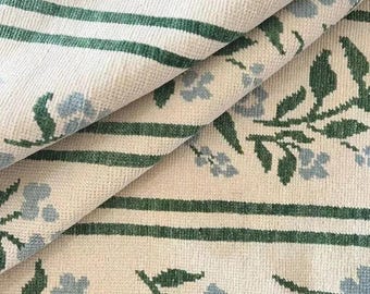 May include: Cream-colored fabric with a repeating pattern of light blue flowers and green leaves. Two dark green horizontal stripes border the floral design. The fabric is folded, showing the texture and pattern.