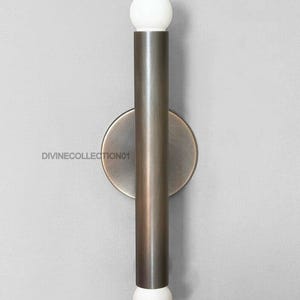 Puede incluir: Aplique de pared moderno con un cuerpo cilíndrico de metal color bronce y una placa trasera circular. La lámpara tiene una bombilla esférica blanca en la parte superior e inferior. El texto "DIVINECOLLECTION01" está impreso en la placa trasera.