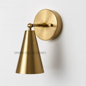 May include: A brass wall sconce with a conical shade and a circular backplate. The sconce has a pivoting arm and is mounted on a white wall. The text "DIVINE COLLECTION01" is visible on the shade.