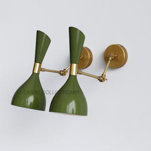 Op de afbeelding: Twee olijfgroene wandlampen met messing accenten. De lampen hebben een uniek, gebogen ontwerp met een uitlopende kap en een cilindrische bovenkant. De tekst "DIVINE COLLECTION01" is zichtbaar.