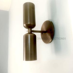 May include: A bronze-colored wall sconce with a cylindrical design. The sconce has two stacked, elongated cylinders connected by a short arm to a circular wall plate. The text "DIVINECOLLECTION01" is visible on the wall.