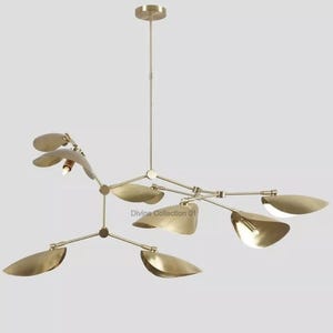 Op de afbeelding: Een moderne messing kroonluchter met bladvormige kappen. De lamp heeft meerdere armen die uitgaan van een centraal punt, elk met een bladvormige kap. De tekst "Divine Collection 01" is zichtbaar.