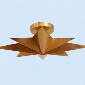 May include: A brass-coloured ceiling light fitting with a starburst design. The fitting has a circular base and multiple triangular points extending outwards. The light is against a light blue background.