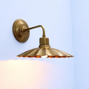 Puede incluir: Un aplique de pared con una placa circular de latón y un brazo curvo que sostiene una pantalla ancha de metal festoneado en tono latón. La lámpara incluye una pequeña bombilla encendida situada bajo la pantalla metálica acanalada.