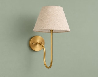 May include: A wall-mounted lamp featuring a circular brass-toned backplate and a curved arm that supports a conical, textured beige fabric lampshade. The fixture has a metallic finish and a simple, functional design for interior lighting.