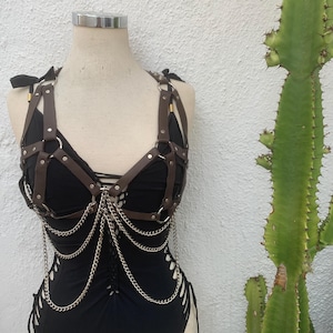 May include: A black bodysuit with brown leather harness detailing and silver chain accents. The harness features a halter neck and connects to the bodysuit with metal rings. The bodysuit has cut-out sides and a lace-up front.