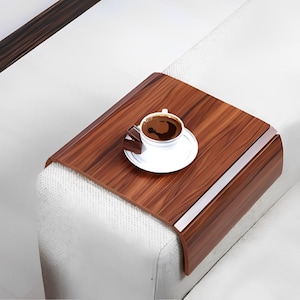 Op de afbeelding: Een bruine houten sofa-armtafel met een glanzende afwerking. Een witte kop en schotel met koffie en een stuk chocolade staan op de tafel. De tafel is ontworpen om over de arm van een bank te passen.