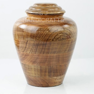 May include: A wooden urn with a lid. The urn is a light brown color with a wood grain pattern. The lid is a darker brown color and has a rounded top. The text "HANDICRAFTS" is visible on the urn.