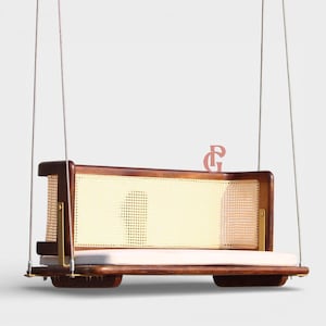 Op de afbeelding: Een houten schommel met een lichtgekleurde geweven rugleuning en zitkussen. De schommel heeft een donkerbruin frame met messingkleurige hardware en is opgehangen aan zilverkleurige touwen. Een roze letter "G" is rechtsboven zichtbaar.