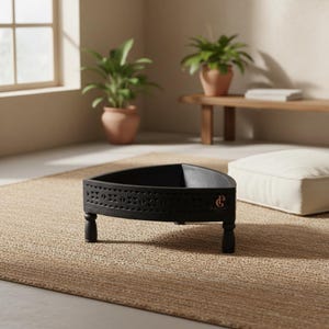 May include: A black, triangular decorative tray with carved geometric patterns along the sides. The tray rests on three short, rounded legs. A small, copper-coloured emblem is visible on the side. The tray is placed on a woven rug.