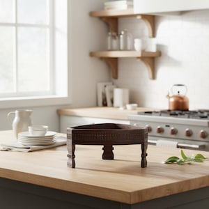 Puede incluir: Una bandeja de madera triangular de color marrón oscuro con detalles tallados. La bandeja tiene patas cortas y decorativas y se exhibe sobre una encimera de madera de color claro en una cocina. La bandeja está vacía.