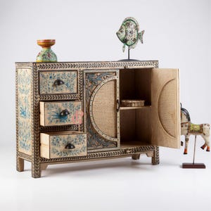 Puede incluir: Un mueble de madera pintado a mano con tres cajones y una puerta, con diseños florales azules y un panel de puerta de caña tejida. El mueble está adornado con detalles decorativos, incluyendo una escultura de pez y una pequeña figura de caballo.