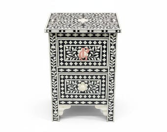 May include: Black and white floral patterned nightstand with two drawers. The nightstand features a decorative floral design on the top, sides, and drawer fronts. The drawer pulls are a light pink color. The nightstand is supported by four small feet.