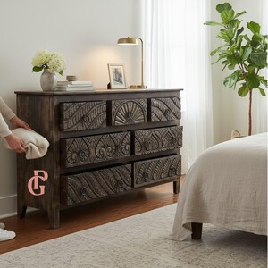 May include: A dark brown wooden dresser with six drawers, each intricately carved with floral and geometric patterns. A gold desk lamp and framed photo sit on top. A person is holding a white blanket next to the dresser.